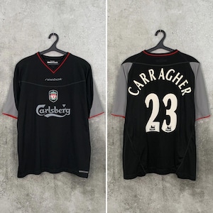 May include: A black Liverpool FC soccer jersey with grey sleeves and red trim. The front features the Reebok logo, the Liverpool crest, and the Carlsberg sponsor text. The back displays the name Carragher and the number 23 in white lettering.