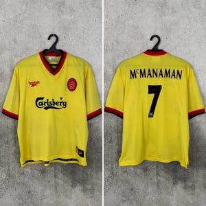 May include: A yellow vintage soccer jersey with red and black trim. The front features the Carlsberg logo, a team emblem, and the Reebok logo. The back displays the name "McManaman" and the number "7".