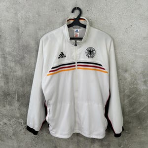 Könnte beinhalten: Eine weiße Adidas Trainingsjacke mit durchgehendem Frontreißverschluss, Stehkragen und langen Ärmeln mit schwarzen Bündchen. Auf der Brust befinden sich ein schwarzes Adidas-Logo und ein rundes deutsches Fußballwappen mit Querstreifen.