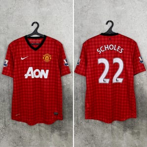 May include: A red Manchester United soccer jersey with a checkered pattern, featuring a white Nike swoosh, the Aon logo, and the Manchester United crest on the front. The back displays the name SCHOLES and the number 22 in white lettering.