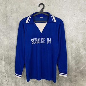 May include: A long-sleeved blue jersey with a white collar and cuffs, hanging on a black hanger. The text "SCHALKE 04" is printed across the chest in white. The jersey has a V-neck design with a white insert.