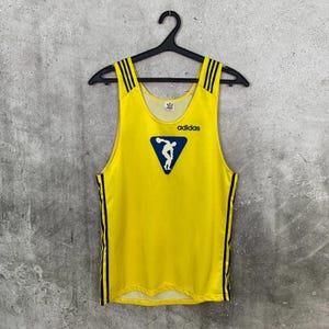 May include: A bright yellow sleeveless tank top featuring dark blue vertical stripes on the shoulders and sides. The chest displays a dark blue triangular graphic of a discus thrower above the word adidas in dark blue text.