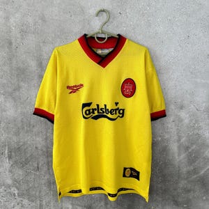 May include: A yellow vintage soccer jersey with red and black trim. The jersey features the Reebok logo, the Carlsberg logo, and a team crest. The jersey is hanging on a hanger against a gray background.