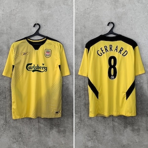 May include: A yellow short-sleeve jersey with black accents, featuring the text Carlsberg on the front and the name GERRARD with the number 8 on the back. The front includes a Reebok logo and a Liverpool football club crest on the chest.