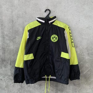 Può includere: Una giacca a vento Nike nera e giallo neon con il logo BVB 09 sul petto. La giacca presenta una chiusura frontale con zip, un colletto alto, maniche lunghe con polsini neri e un orlo regolabile con cordino giallo neon.