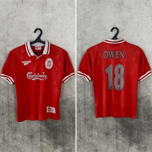 May include: A red short-sleeved jersey with a white collar and sleeve trim. The front features the Reebok logo, a Liverpool crest, and the Carlsberg text. The back displays the name OWEN and the number 18 in grey lettering on a red background.