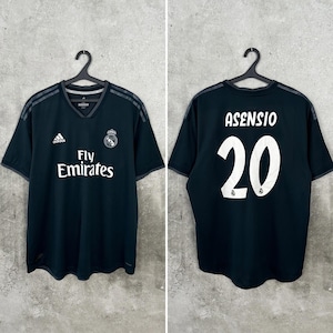 Könnte beinhalten: Ein schwarzes Real Madrid Fußballtrikot mit dem Adidas-Logo, dem Fly Emirates Sponsorentext und dem Vereinswappen auf der Vorderseite. Auf der Rückseite stehen der Name ASENSIO und die Nummer 20 in weißer Schrift. Das Trikot hat kurze Ärmel und einen V-Ausschnitt.