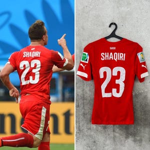 May include: A red football shirt featuring the name SHAQIRI and the number 23 in white on the back. The shirt includes the word SUISSE, white Puma logos on the sleeves, and two circular patches on the shoulders, all displayed on a black hanger.
