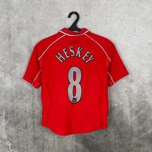 May include: A red short-sleeved soccer jersey with the name "HESKEY" and the number "8" in silver lettering. The jersey has white trim around the collar and sleeves. A black hanger holds the jersey against a gray background.