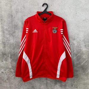 May include: A bright red Adidas track jacket featuring a full-length front zip, a high collar, and long sleeves with three white stripes running down each arm. The jacket displays a white Adidas logo and a crest with an eagle emblem on the chest.