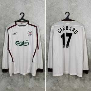 May include: White long-sleeved jersey with red and black accents. The front features the Liverpool crest, the Carlsberg logo, and the Reebok logo. The back displays "GERRARD" above the number "17".