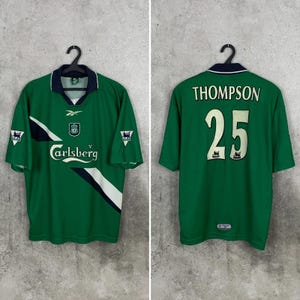 May include: Green soccer jersey with white and black accents, featuring the Carlsberg logo and the number 25 with the name "THOMPSON" on the back. The jersey has a V-neck collar and short sleeves. The jersey is hanging on a black hanger.