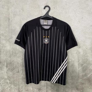 Könnte beinhalten: Ein schwarzes Kurzarmtrikot mit dünnen vertikalen weißen Nadelstreifen, einem Rundhalsausschnitt und drei goldenen Sternen über einem kreisförmigen Emblem auf der Brust. Unten rechts befinden sich drei diagonale weiße Streifen und das Wort Deutschland.