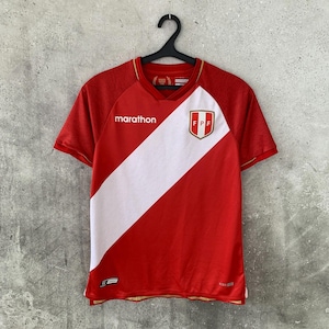 Peut inclure: Un maillot à manches courtes rouge et blanc avec une bande blanche diagonale sur le devant, le mot marathon en texte blanc et l'écusson FPF sur la poitrine. Le maillot possède un col en V et est présenté sur un cintre en plastique noir.