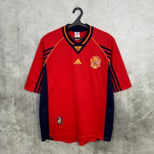 May include: A red short-sleeved soccer jersey featuring dark blue side panels and three dark blue stripes on the shoulders. The shirt displays a yellow Adidas logo, the Spanish national football crest, and a small RFEF emblem on the front.