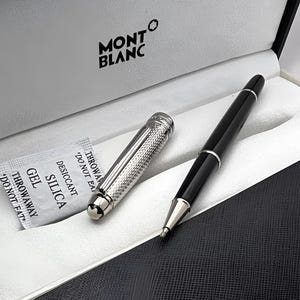 Può includere: Una penna a sfera Montblanc nera con cappuccio e dettagli argentati, presentata in una scatola bianca. La penna è accompagnata da una bustina di essiccante e un taccuino nero. Il logo Montblanc è visibile.