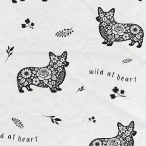 May include: White fabric featuring a repeating pattern of black corgi dog silhouettes filled with floral designs. The phrase "wild at heart" is written in black script, along with small floral and leaf accents.