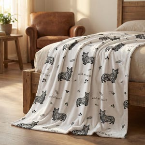 May include: A white blanket with a repeating pattern of black corgi dog illustrations and the text "wild at heart". The blanket is draped over a wooden bed frame, creating a cozy and decorative look.