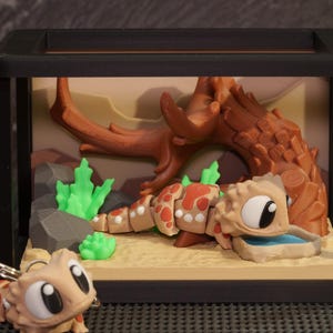 May include: A miniature terrarium scene featuring a cartoon lizard with a red and tan pattern, a water dish, and a miniature keychain version. The background includes a brown tree branch, green plants, and a sandy base, all within a black framed enclosure.