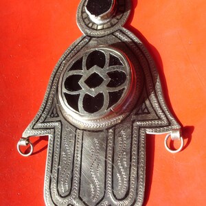 May include: A silver charm in the shape of a hamsa hand with black onyx stones. The charm has intricate details and a loop at the top for hanging.