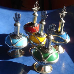 Pode incluir: Cinco garrafas decorativas com tampas prateadas e bases de vidro coloridas. As garrafas têm um design em forma de mão na parte superior e estão dispostas sobre uma superfície azul.