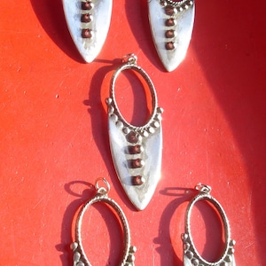 May include: Five silver pendant earrings with a teardrop shape and a circular opening. Each earring has a row of small silver beads around the edge and four dark red beads in the center.