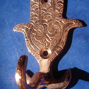 Peut inclure: Crochet mural en cuivre en forme de main de hamsa avec des détails complexes. Le crochet a deux pointes pour accrocher des objets.