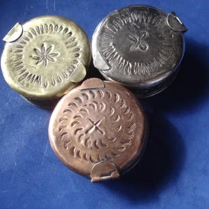 May include: Three small, round metal containers with hinged lids. One is brass-colored, one is silver-colored, and one is copper-colored. Each has a decorative, embossed design on the lid. The brass container has a star design, the silver one has a flower, and the copper one has a sunburst.