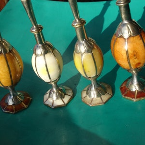 Puede incluir: Cuatro botellas decorativas con tapas plateadas y cuerpos de vidrio ámbar, blanco y marrón. Las botellas tienen diseños intrincados y están dispuestas en una fila sobre una superficie turquesa.