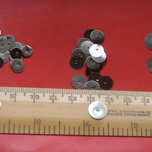 May include: A close-up of three piles of silver metal sequins. The sequins are different sizes and are arranged in a random pattern. A ruler is placed below the sequins for size reference.
