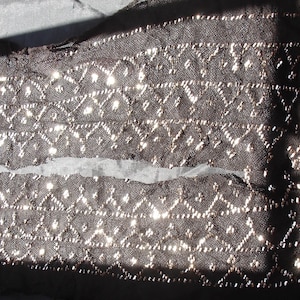 Puede incluir: Tela de encaje marrón oscuro con un patrón de diamantes y detalles metálicos. El encaje está parcialmente rasgado, revelando una tela más clara debajo. La tela probablemente se utiliza para ropa o fines decorativos.