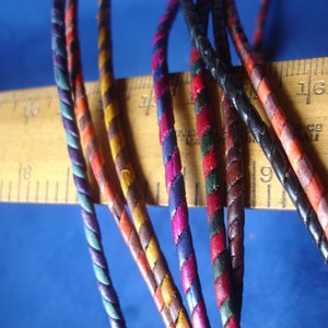 May include: Close-up of several colourful braided leather cords. The cords are in various colours, including red, orange, yellow, green, blue, and black. A wooden ruler is in the background. The cords are likely used for jewellery making or crafts.