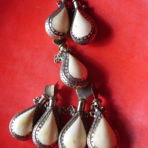 May include: A collection of six teardrop-shaped containers featuring smooth, off-white, rounded centres encased in ornate, textured silver-toned metal frames. Each piece includes a small metal stopper attached by a delicate chain to the main body.