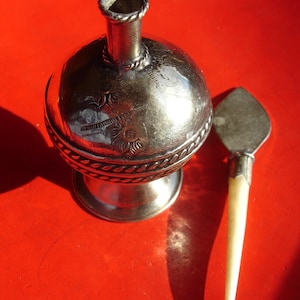 May include: A silver, ornate, round bottle with a small, silver, pointed applicator. The bottle has a decorative design and a small, round base. The applicator is long and slender.