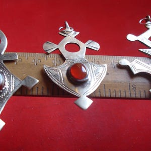 May include: Three silver pendants with geometric designs and gemstone accents. Each pendant features a different shape, with a central gemstone in shades of orange and brown. The pendants are displayed on a wooden ruler against a red background.