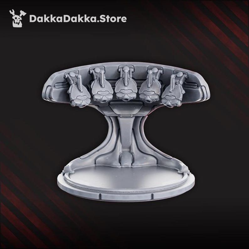 May include: A gray, futuristic-looking stand with five detailed, stylized objects on top. The stand has a wide, circular base and a central support that flares out to hold the top section. The text "DakkaDakka.Store" is in the upper left corner.