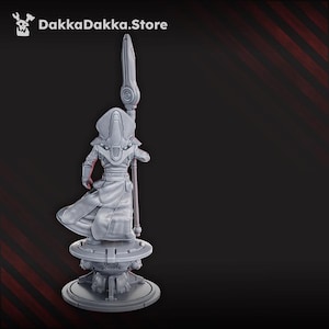 May include: A gray figurine of a robed figure holding a staff with a detailed headpiece, standing on a circular platform. The figurine is set against a dark background with diagonal red stripes. The text "DakkaDakka.Store" is in the top left corner.