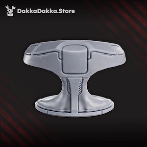 May include: A gray, futuristic-looking object with a wide, flat top and a curved base. The top has a series of rectangular panels and a central, raised section. The base is rounded and supported by a stylized, hourglass-shaped stem. The text "DakkaDakka.Store" is in the top left corner.