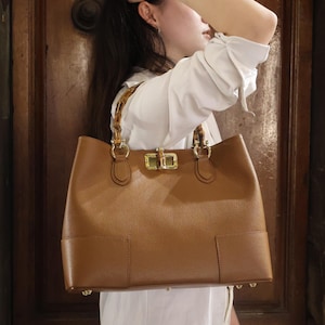 Può includere: Una grande borsa tote in pelle marrone con manici in bambù e finiture color oro. La borsa ha una forma rettangolare con un design strutturato e sembra essere realizzata con materiali di alta qualità. La borsa è un accessorio elegante.