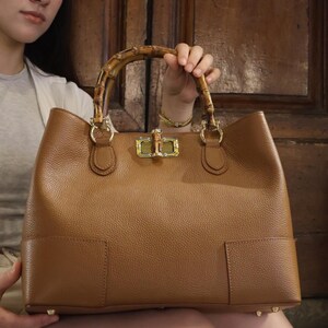 May include: A tan leather handbag with a textured surface, featuring a curved bamboo handle and a gold-toned metal turn-lock closure. The bag has a structured rectangular base with visible stitching details and metallic feet on the bottom corners.