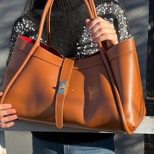 K&ouml;nnte beinhalten: Eine gro&szlig;e, braune Ledertasche mit rotem Innenfutter. Die Tasche hat zwei Griffe und einen silberfarbenen Schnallenverschluss. Die Tasche wird von einer Person gehalten.