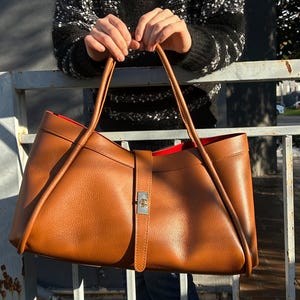 K&ouml;nnte beinhalten: Eine gro&szlig;e, braune Ledertasche mit rotem Innenfutter. Die Tasche hat lange Henkel und einen Riemenverschluss mit silberfarbener Schnalle. Die Tasche wird von einer Person gehalten, die einen schwarz-wei&szlig; gemusterten Pullover tr&auml;gt.