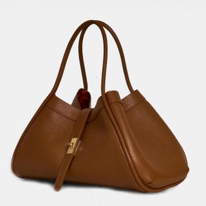 K&ouml;nnte beinhalten: Eine braune Ledertasche mit einer einzigartigen dreieckigen Form. Die Tasche hat zwei lange Henkel, einen goldfarbenen Verschluss und einen Riemenverschluss. Das Innere ist mit rotem Stoff gef&uuml;ttert. Das Design der Tasche ist minimalistisch.