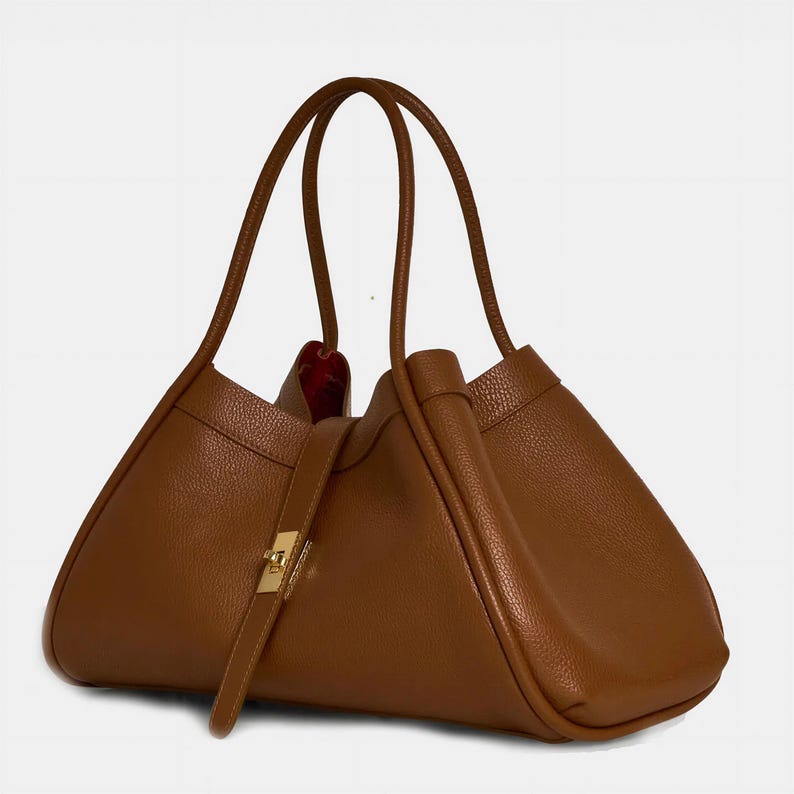 K&ouml;nnte beinhalten: Eine braune Ledertasche mit einer einzigartigen dreieckigen Form. Die Tasche hat zwei lange Henkel, einen goldfarbenen Verschluss und einen Riemenverschluss. Das Innere ist mit rotem Stoff gef&uuml;ttert. Das Design der Tasche ist minimalistisch.