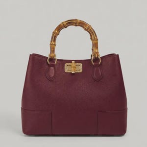 May include: A deep burgundy handbag with a textured surface, featuring a curved bamboo handle and a gold-toned metal turn-lock closure with a small bamboo accent. The bag has a structured base and two vertical rectangular panels at the bottom front.