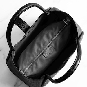 K&ouml;nnte beinhalten: Schwarze Ledertasche mit zwei abgerundeten Griffen. Die Tasche hat eine strukturierte Au&szlig;enseite und ein Innenfach mit Rei&szlig;verschluss. Das Innenfutter ist ein dunkelgrauer Stoff. Die Tasche ist offen und gibt den Blick ins Innere frei.