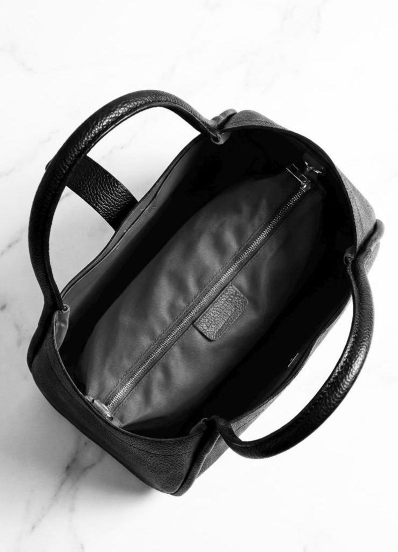 K&ouml;nnte beinhalten: Schwarze Ledertasche mit zwei abgerundeten Griffen. Die Tasche hat eine strukturierte Au&szlig;enseite und ein Innenfach mit Rei&szlig;verschluss. Das Innenfutter ist ein dunkelgrauer Stoff. Die Tasche ist offen und gibt den Blick ins Innere frei.