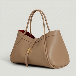 K&ouml;nnte beinhalten: Eine hellbraune Ledertasche mit rotem Innenfutter. Die Tasche hat zwei Henkel und einen Riemen mit goldenem Verschluss. Das Design ist schlicht und elegant, geeignet f&uuml;r den t&auml;glichen Gebrauch.