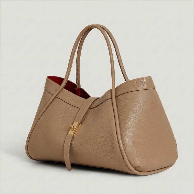K&ouml;nnte beinhalten: Eine hellbraune Ledertasche mit rotem Innenfutter. Die Tasche hat zwei Henkel und einen Riemen mit goldenem Verschluss. Das Design ist schlicht und elegant, geeignet f&uuml;r den t&auml;glichen Gebrauch.