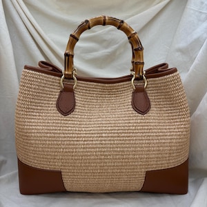 Pode incluir: Uma bolsa tote tecida com uma al&ccedil;a de bambu. A bolsa apresenta um corpo tecido castanho claro com detalhes em couro castanho nos cantos inferiores e nas fixa&ccedil;&otilde;es da al&ccedil;a. A al&ccedil;a &eacute; feita de bambu curvo.